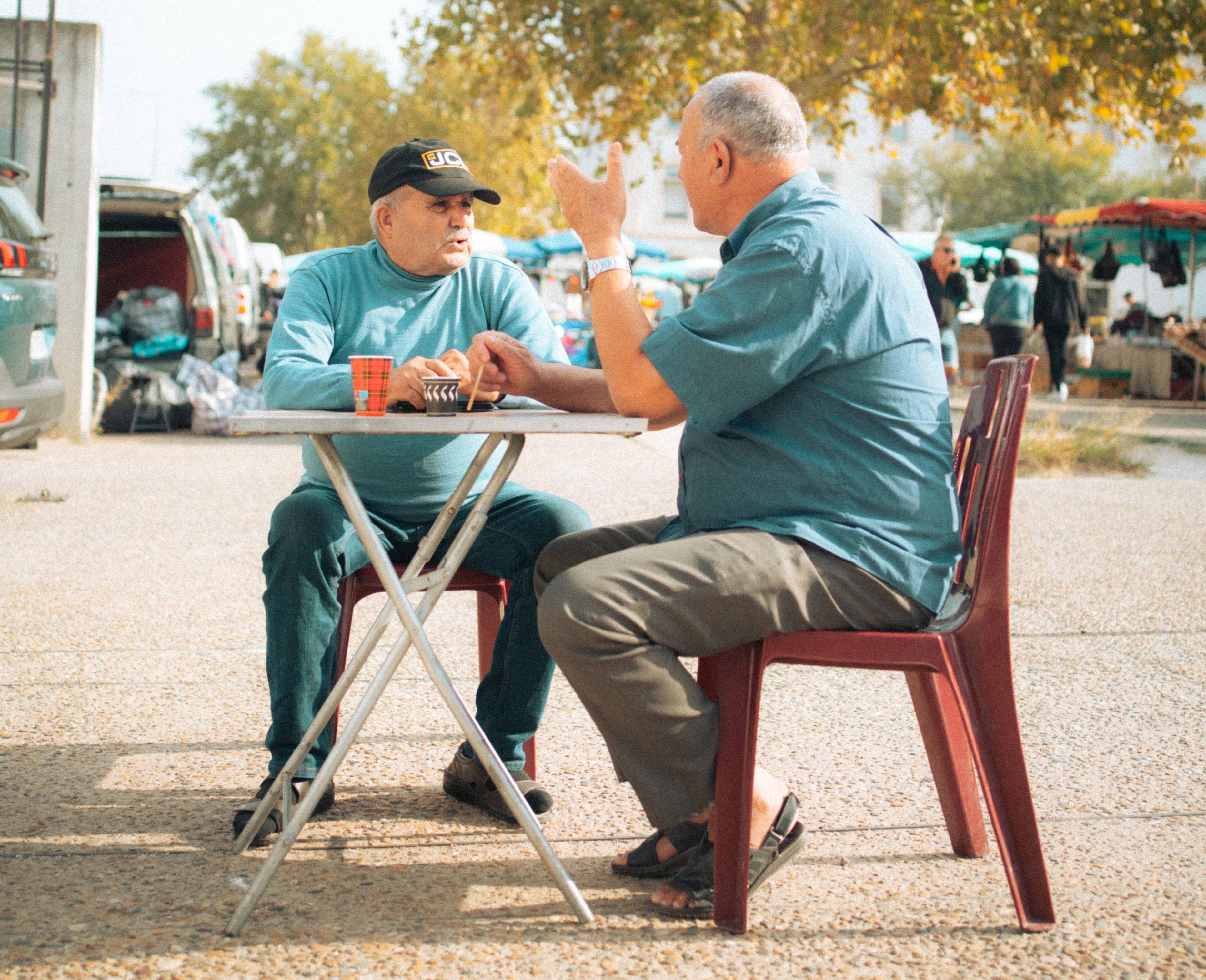 Deux hommes discutant autour d’une table sur un marché populaire, scène de vie captée dans l’exposition Jusqu’à midi de Lola Gadéa