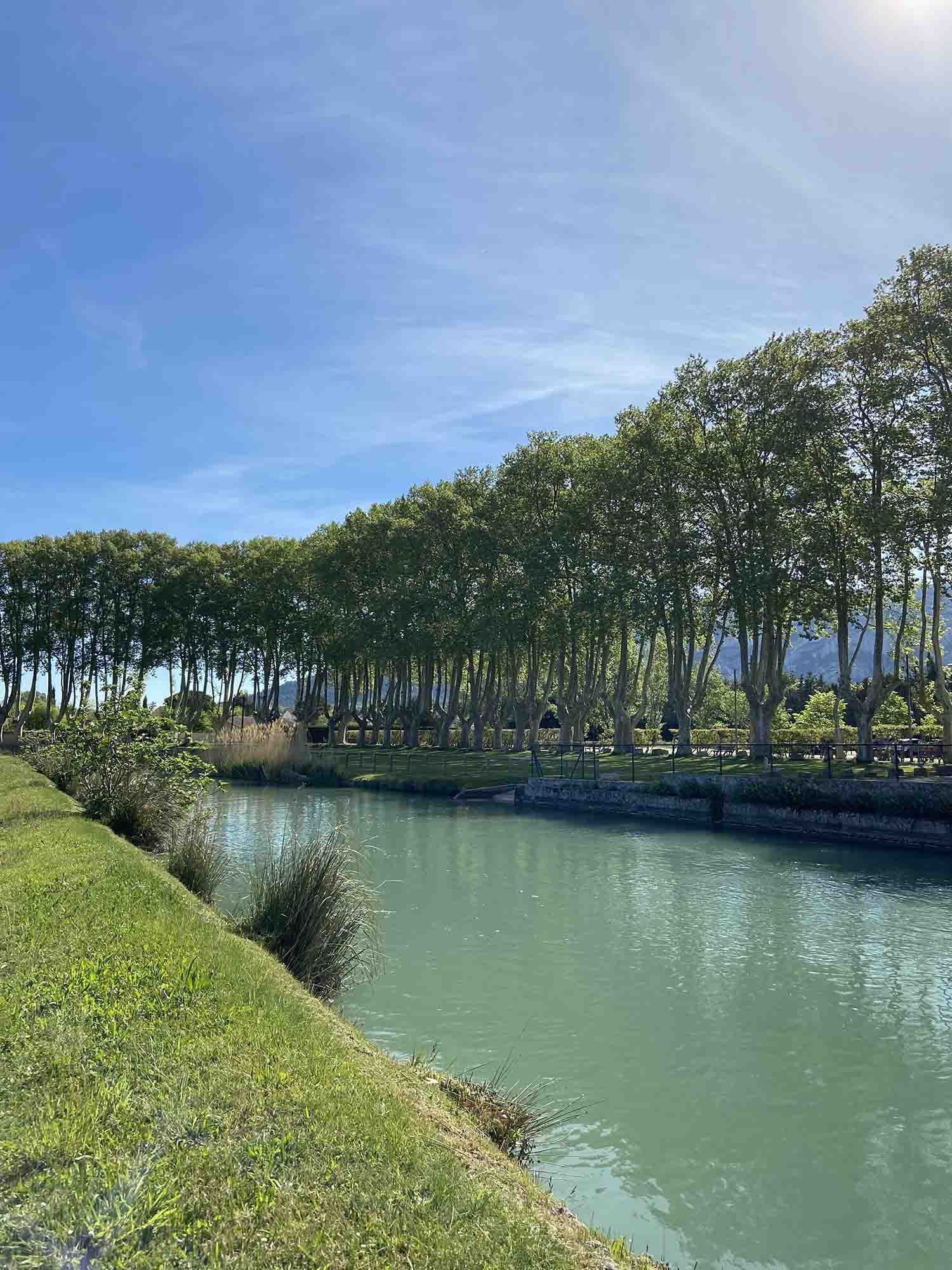 Une pause rafraîchissante au bord de l'eau au Moulin Saint-Pierre Vue du Moulin Saint-Pierre aux Taillades avec sa grande roue à aubes en bois au bord du canal, entouré de verdure et de fleurs sous un ciel bleu.