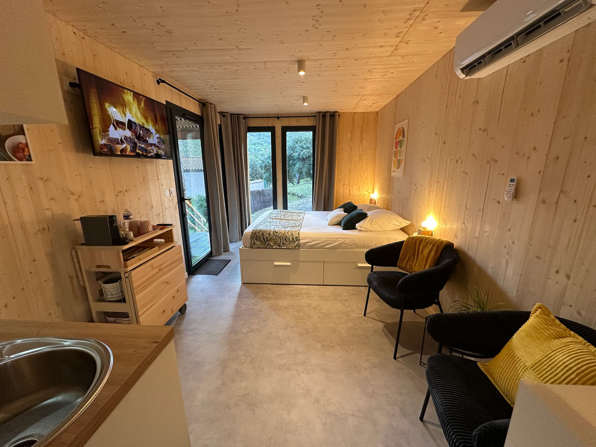 Vue intérieure de la cabane avec lit double, fauteuils, télévision et coin cuisine