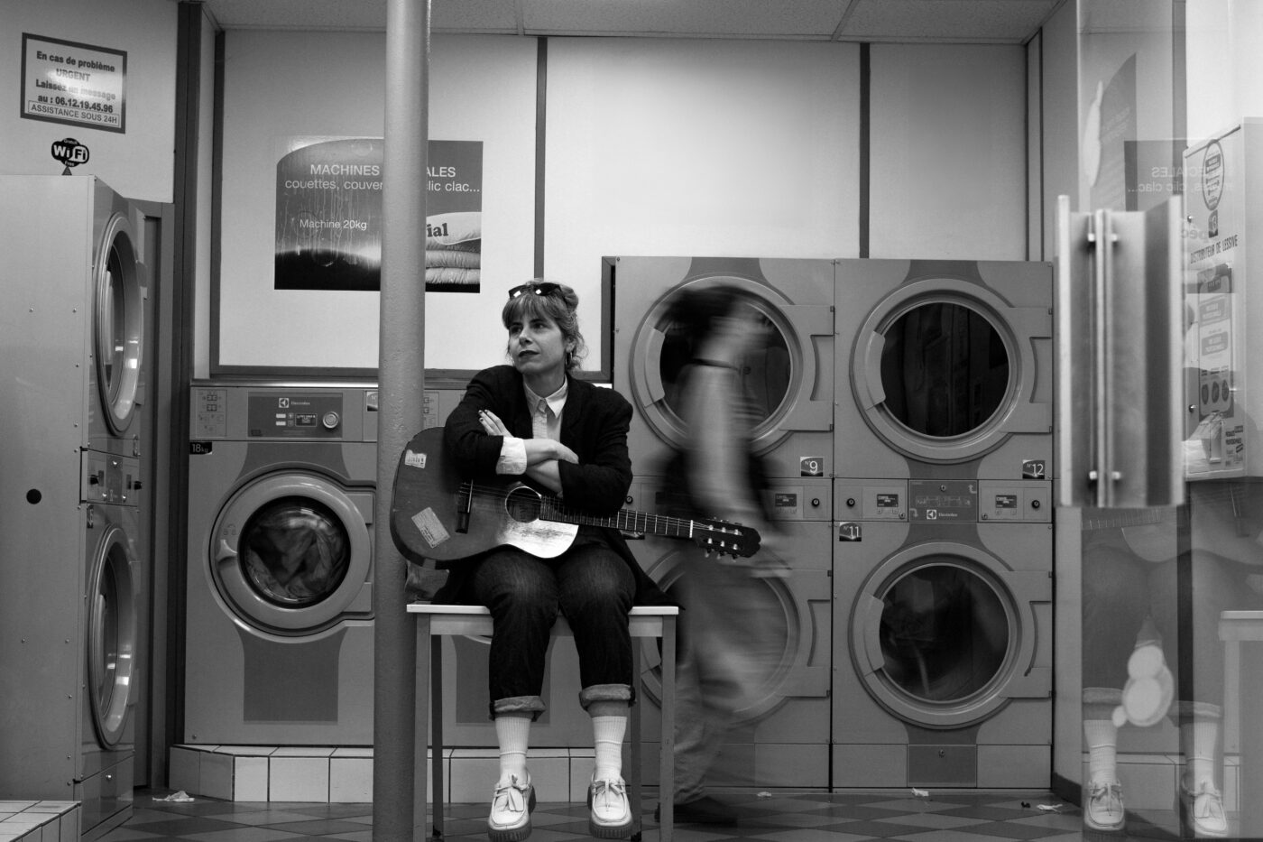 Portrait en noir et blanc de la chanteuse Marina P assise avec une guitare dans une laverie